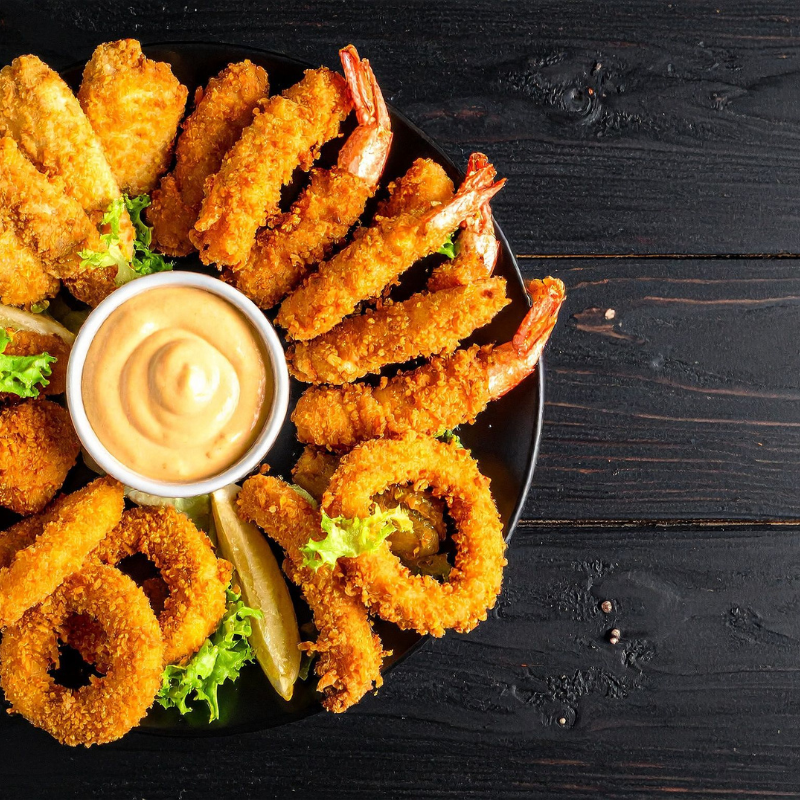 Plate of fried food: chicken, shrimp, onion rings, with dipping sauce on a dark wood table.