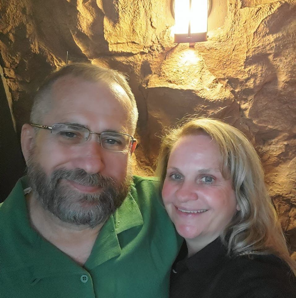 Man with glasses and beard, in green shirt, smiles with woman in black shirt; stone wall backdrop.
