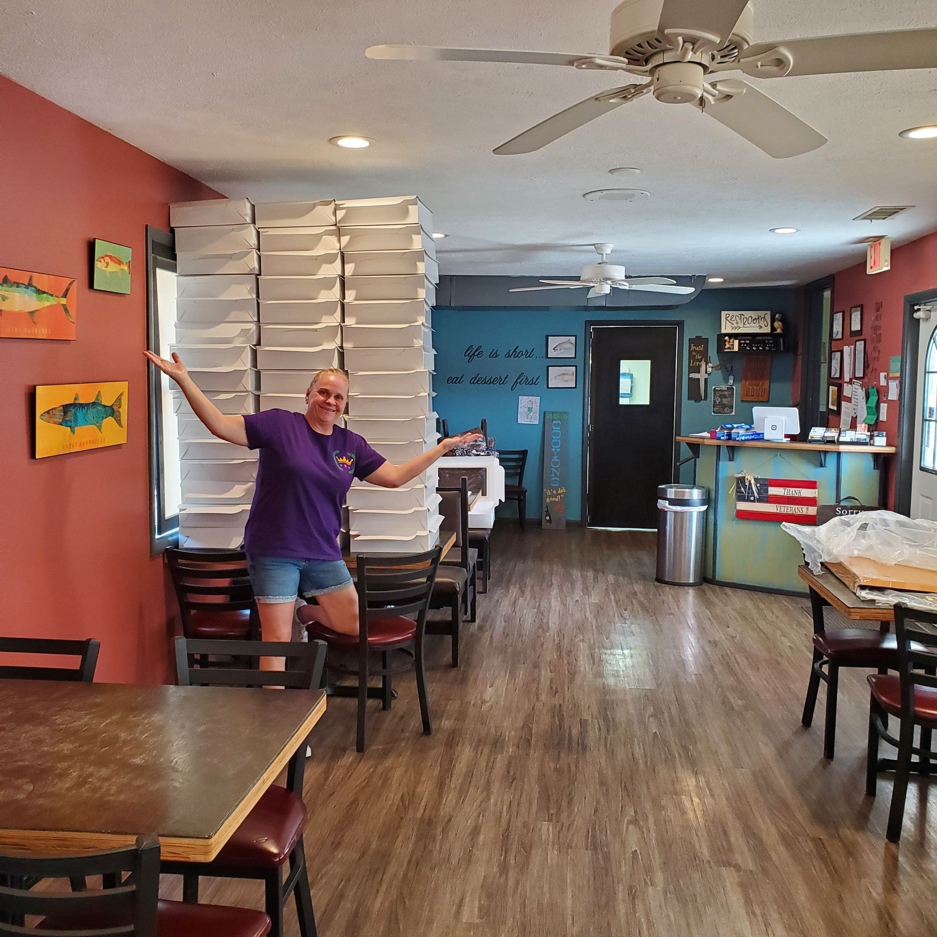 Woman in restaurant smiles, arms wide, by tall stack of pizza boxes.