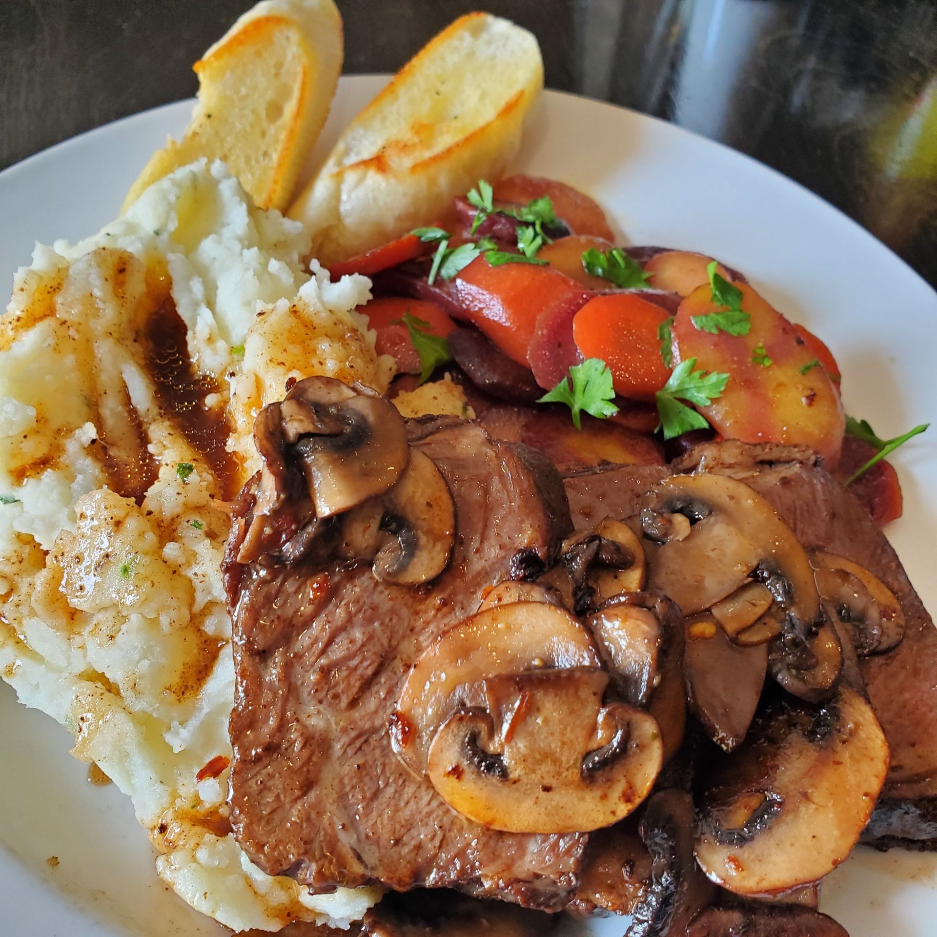 Plate of meat with mushroom sauce, mashed potatoes, glazed carrots, and toasted bread.