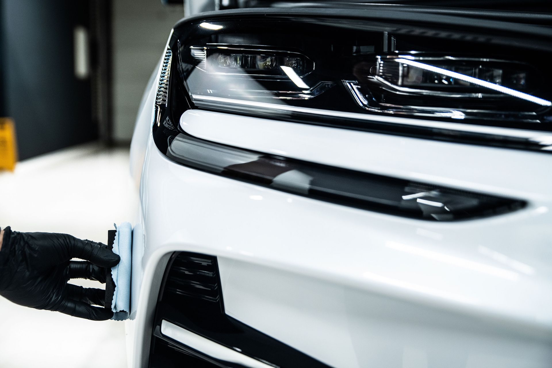 A gloved hand applies coating to a white car's front bumper, near a headlight.