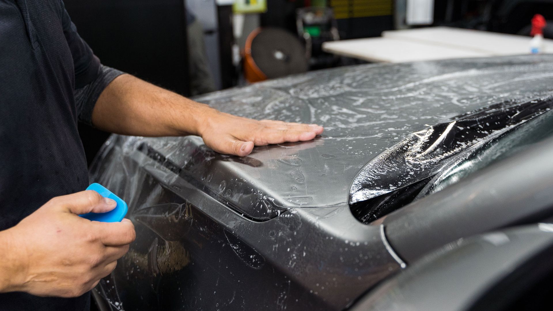 Person applying clear film to a car's hood in a workshop, using a squeegee.