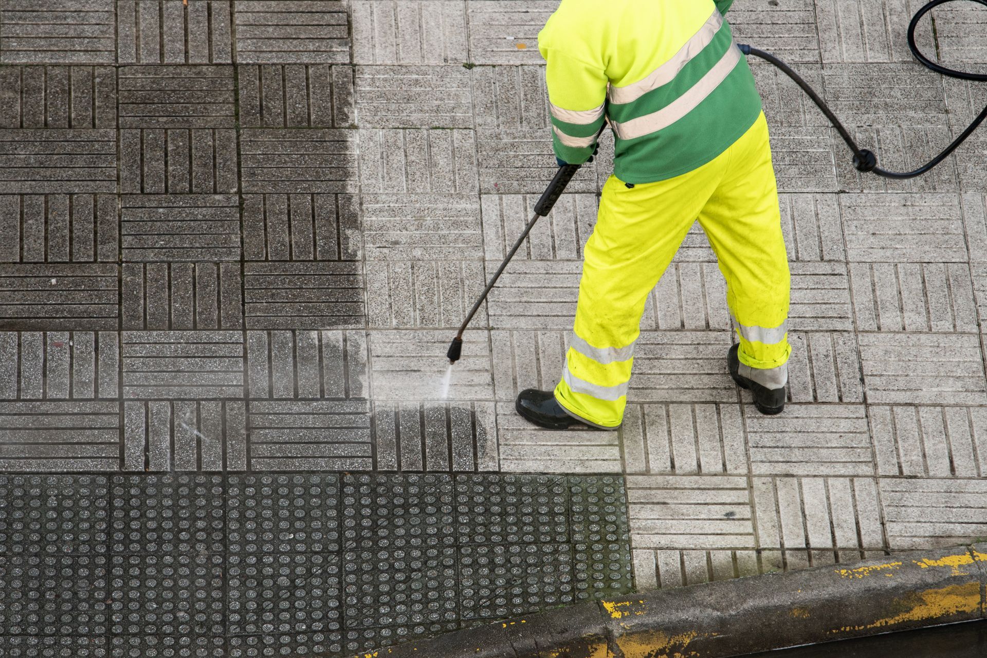 Person in yellow cleaning the sidewalk with a pressure washer; the cleaned portion is noticeably lighter.