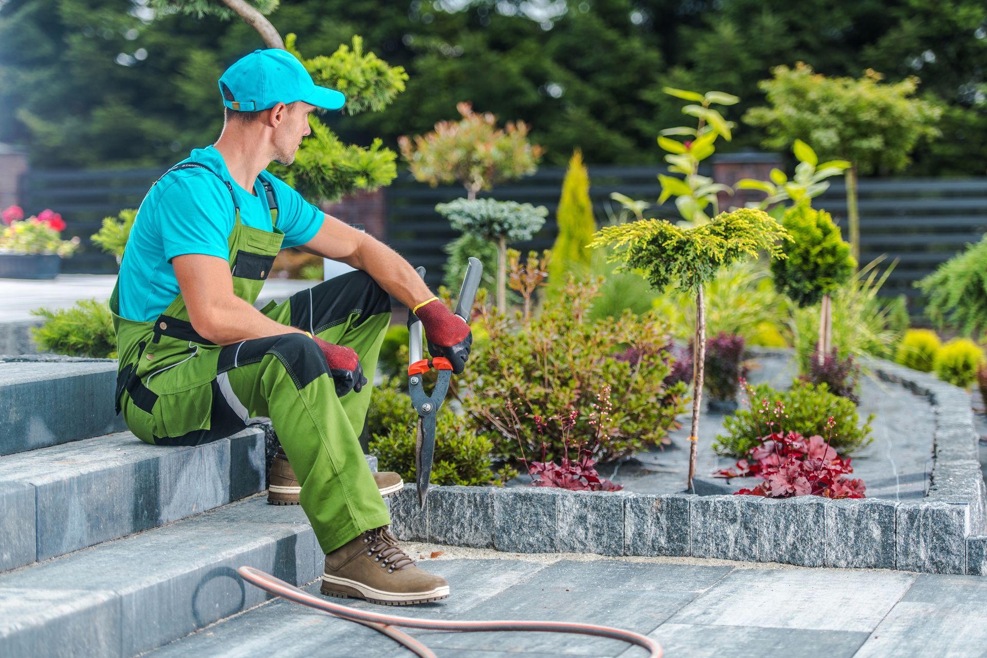 Gardener in green overalls and cap rests on steps, holding pruning shears, overlooking garden beds.