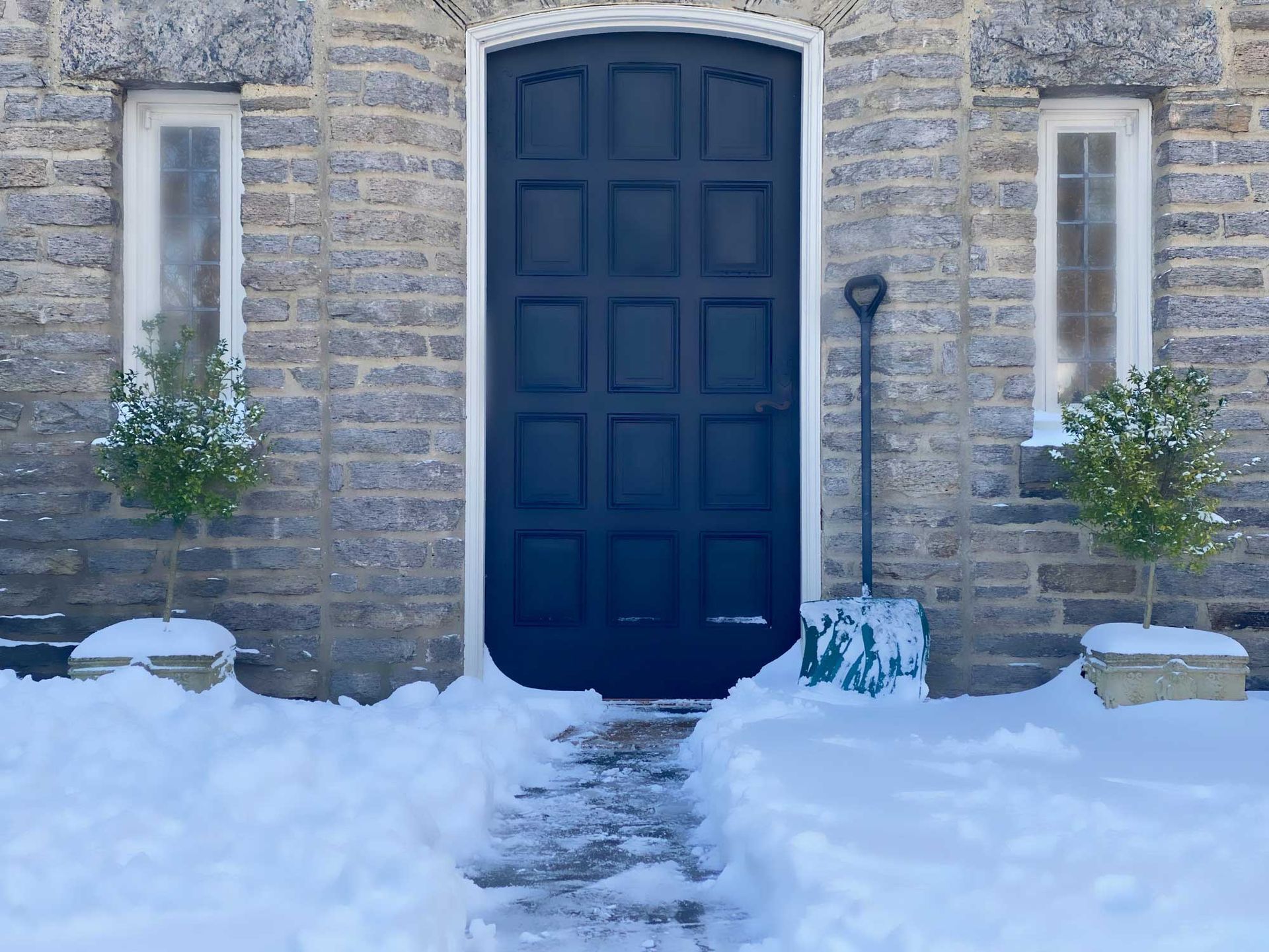 Snow-covered doorway with a dark blue door, flanked by windows and bushes, a shovel leans against the door.