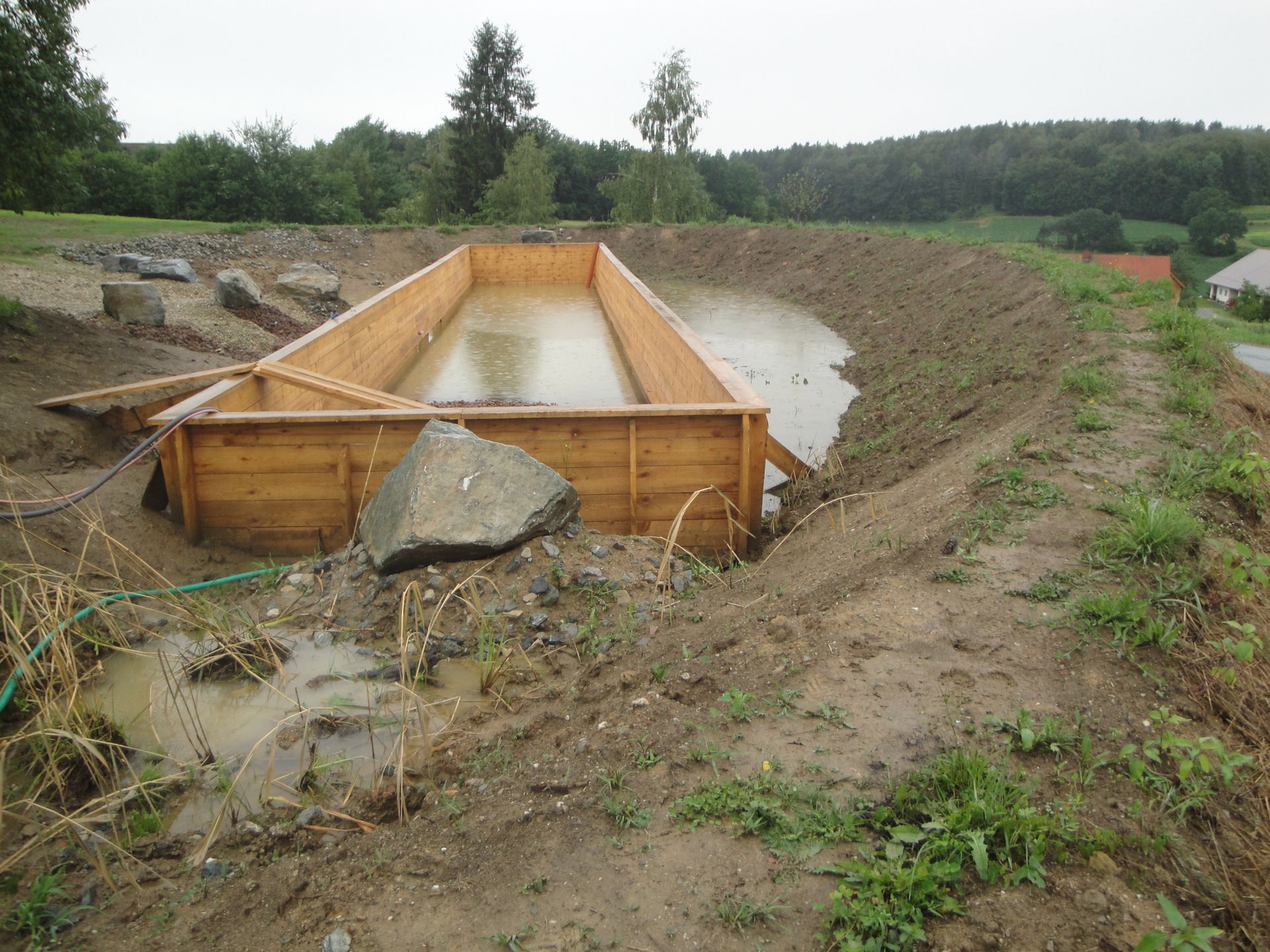 Wooden rectangular structure within a muddy, excavated area, likely a pond under construction.