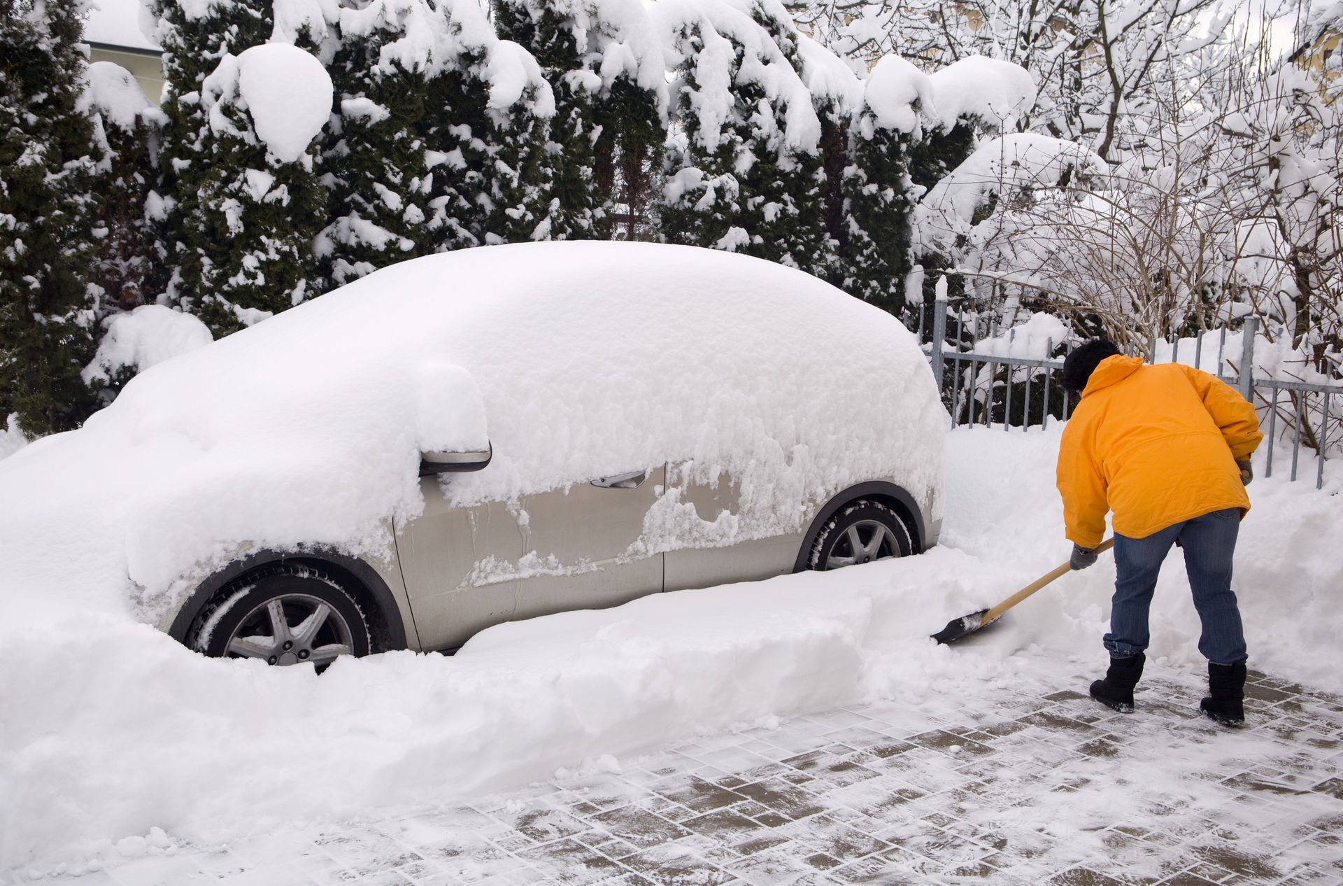 Person shoveling snow from driveway, car covered in snow.