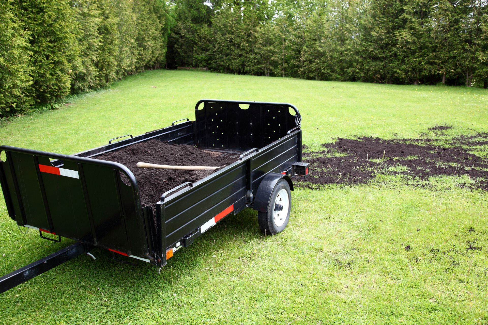 Black trailer filled with soil on a green lawn, near a dark soil spill.