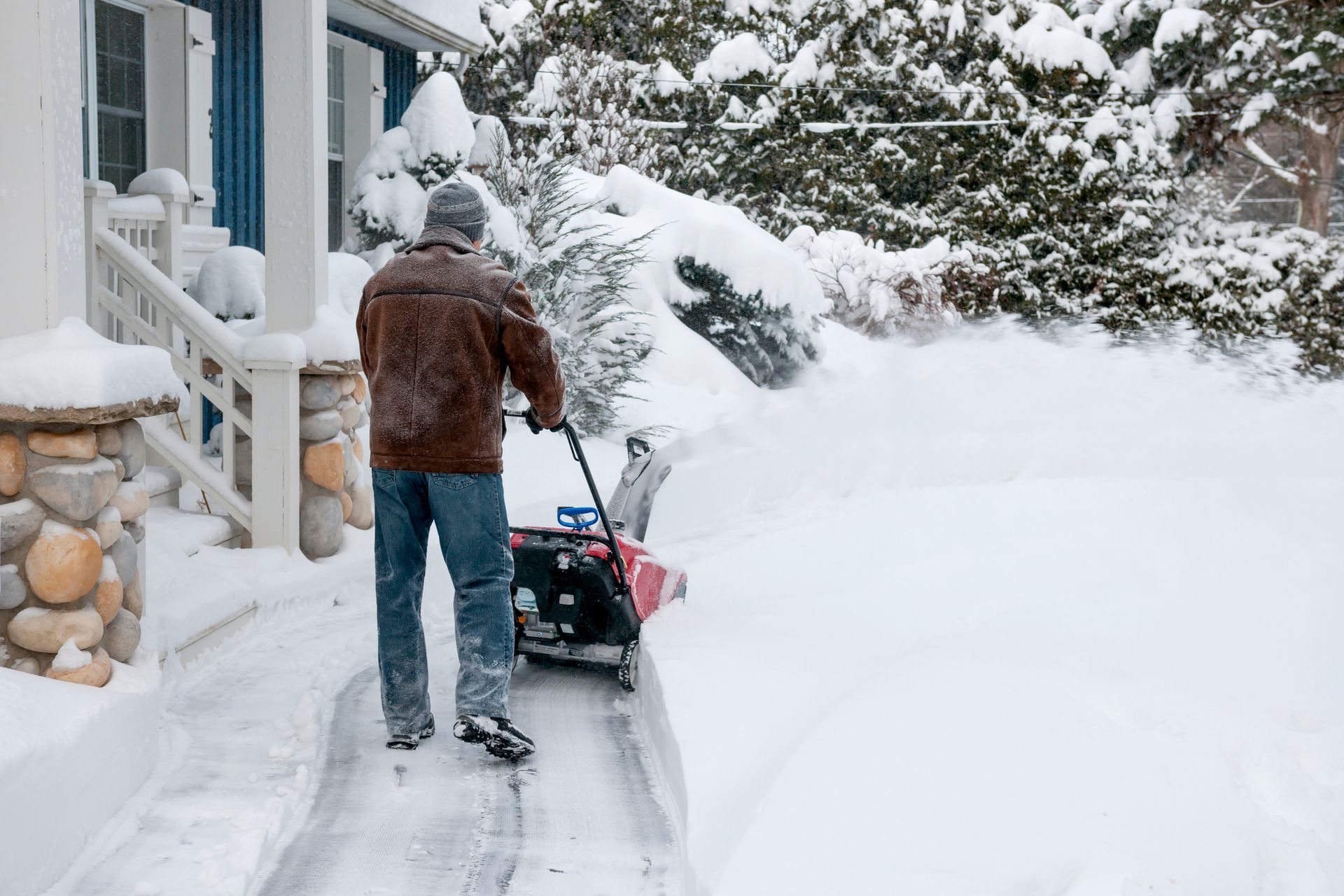 Man uses snowblower on a snowy walkway, clearing a path in front of a house during winter.