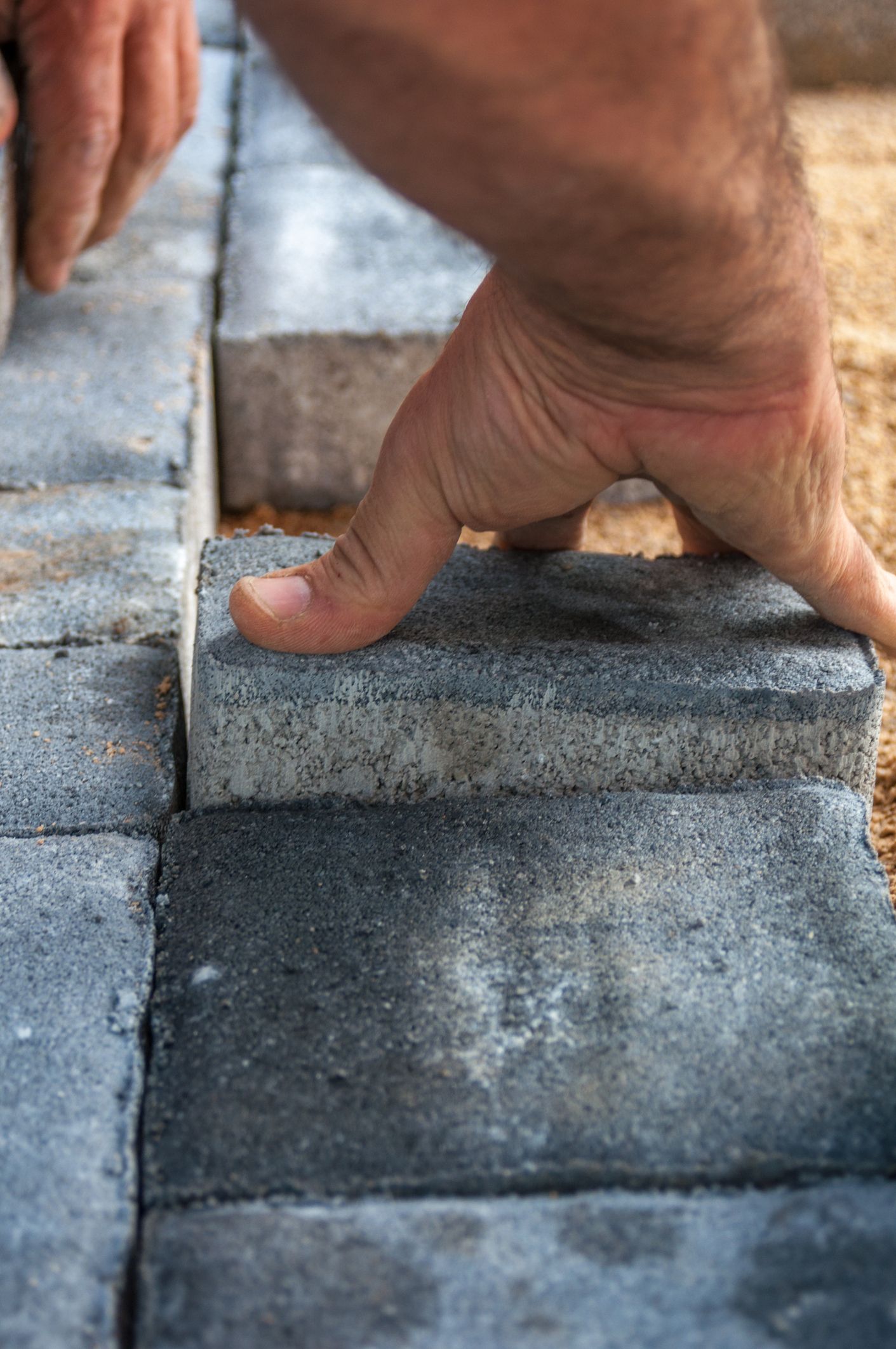 Person's hands placing a gray paver brick onto a paved surface.