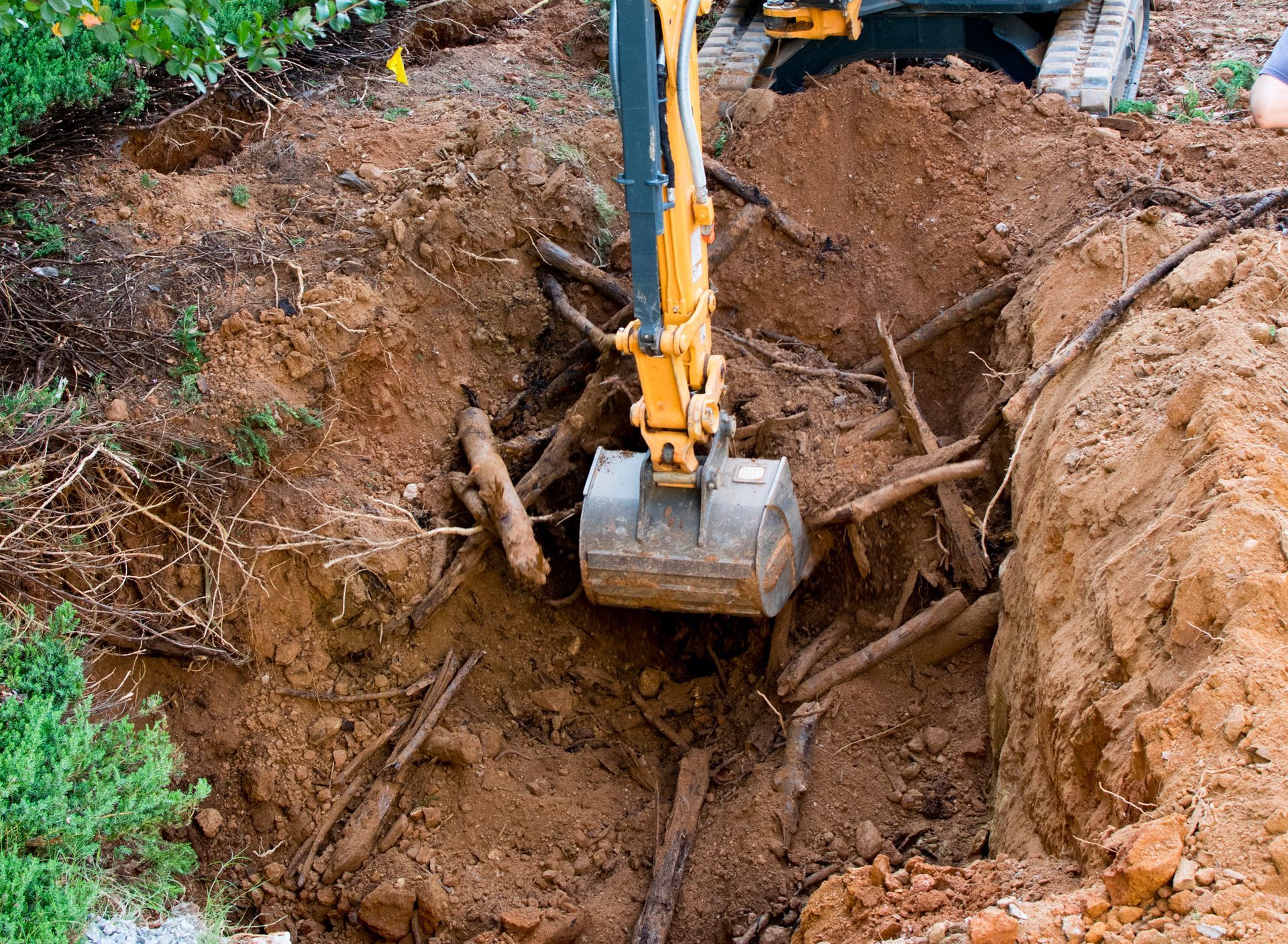 A small excavator digging in a dirt pit, removing wooden branches and roots.