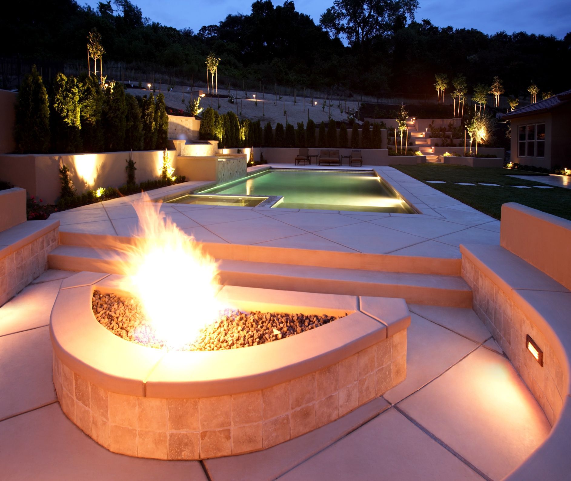 Outdoor patio with a lit fire pit, pool, and landscape lighting at dusk.