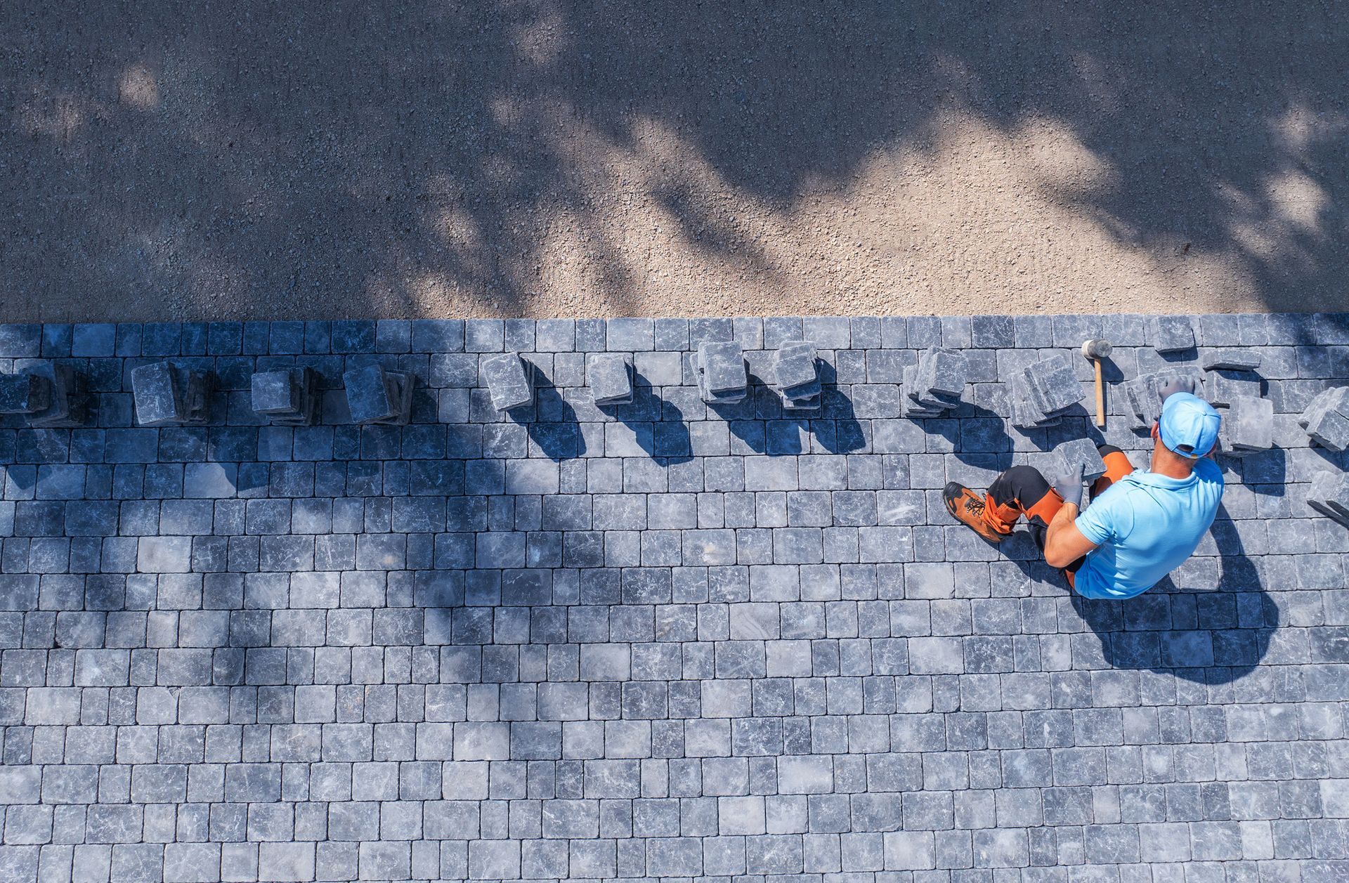 Construction worker laying gray pavers, top view. Bright blue shirt, sunny day.