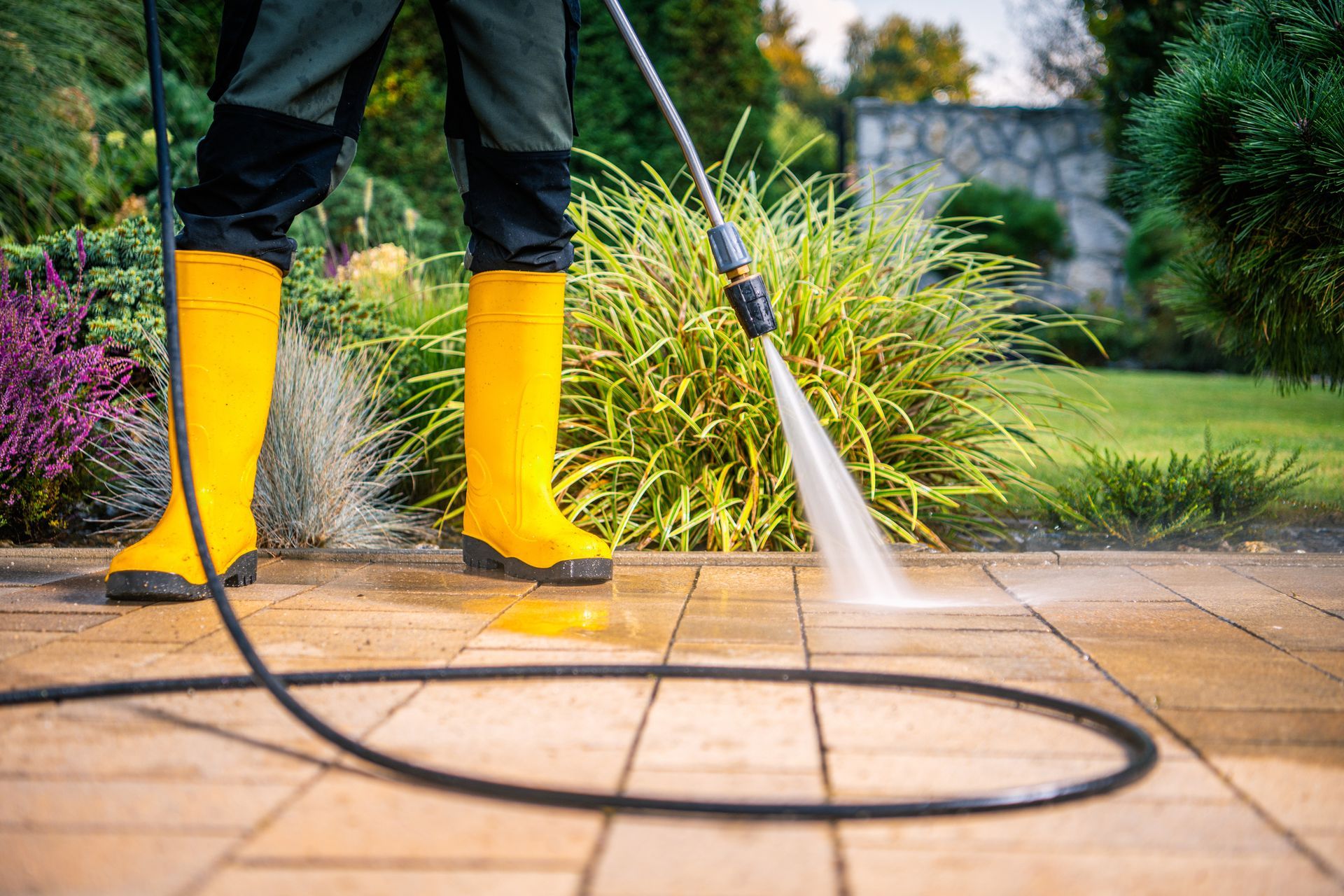 Person power washing a brick patio, wearing yellow boots. Outdoors in a garden setting.