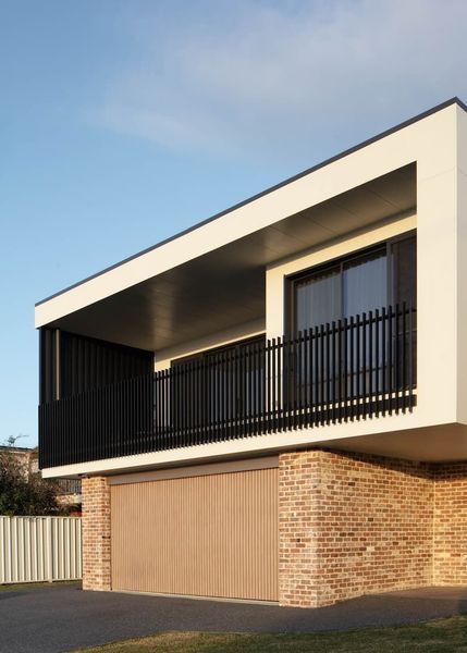 Modern two-story building with a white upper level, black railing, and brick and brown garage door on the ground floor.