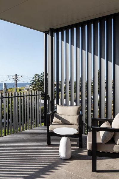 Balcony with two armchairs and a small table. Black vertical slats cast shadows, with a view of the ocean.