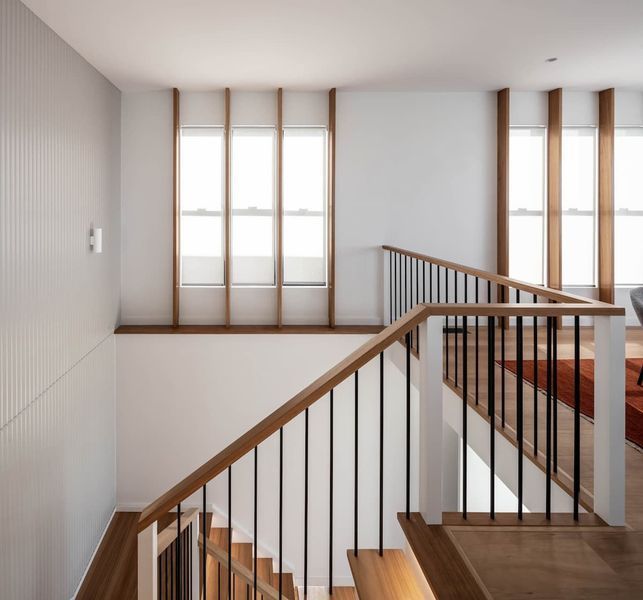 Interior staircase with wood railing and black vertical bars, next to windows with wooden frames.