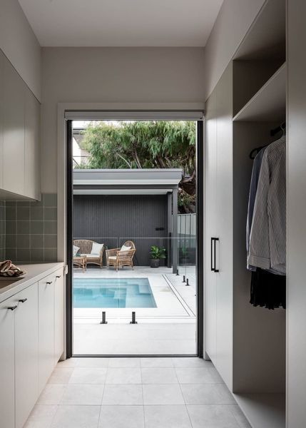 A narrow laundry room opens to a pool area. Light grey cabinetry, white tiles, and a wardrobe with clothes.