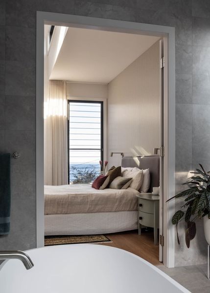A bedroom seen through an open doorway from a bathroom, with a bed, window, and plants.