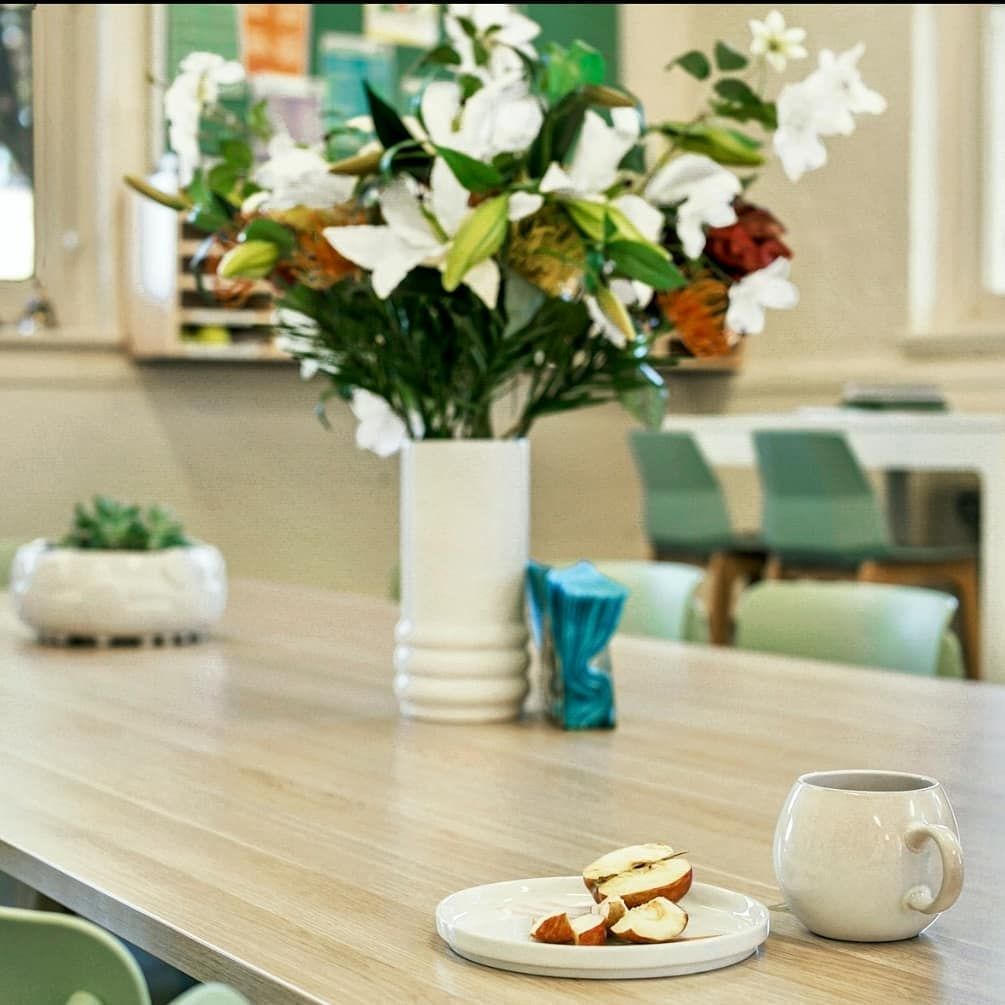 Close-up of table with flower vase, plate with apple slices, and mug. Green chairs and succulents visible.