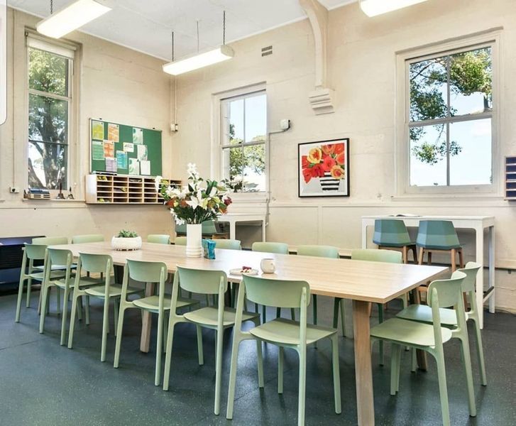 Light green chairs surround a large wooden table in a bright room with windows, plants, and colorful artwork.
