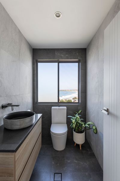 Modern bathroom with gray tiled walls, wooden vanity, black sink, toilet, and plant by a window with a beach view.