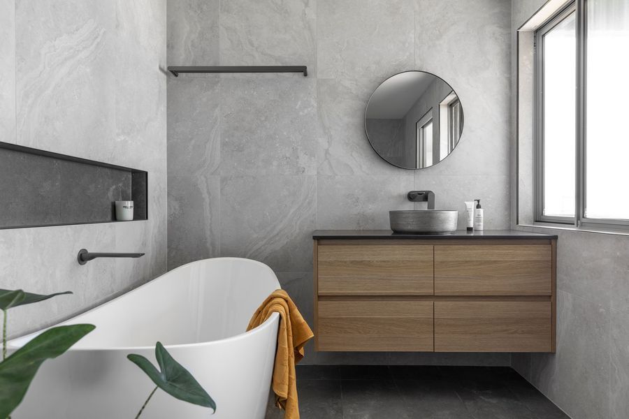 Modern bathroom: a white tub, wooden vanity with a round mirror, and gray textured walls.