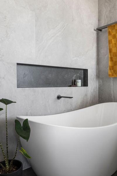 Freestanding white bathtub against a gray tile wall with a recessed shelf and a black faucet.