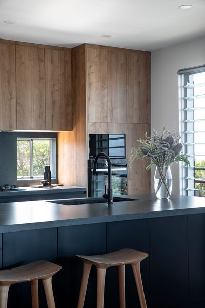 Modern kitchen with wooden cabinets, dark countertops, and bar stools. A vase of flowers sits on the counter.