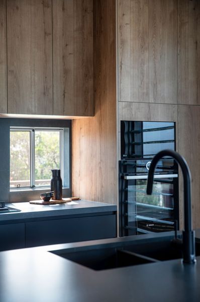 Modern kitchen with wood cabinets, dark countertops, and built-in oven. Black faucet and window.