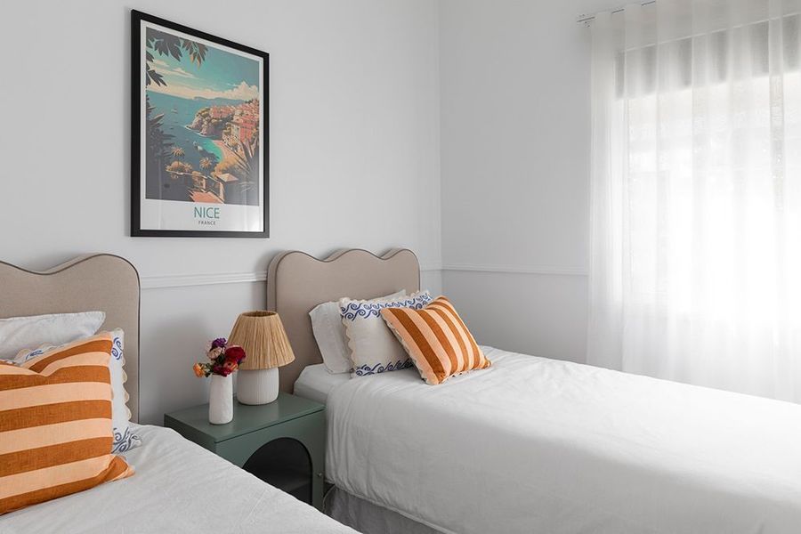 Two beds in a white room with orange striped pillows, green nightstand, and artwork on wall.