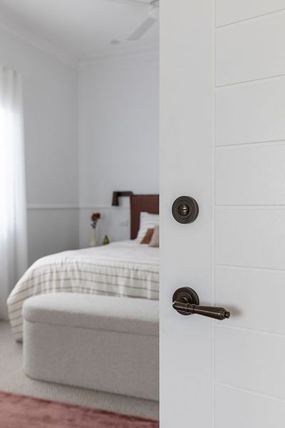 White door partially open to reveal a bedroom with a bed and bench. Bronze hardware.