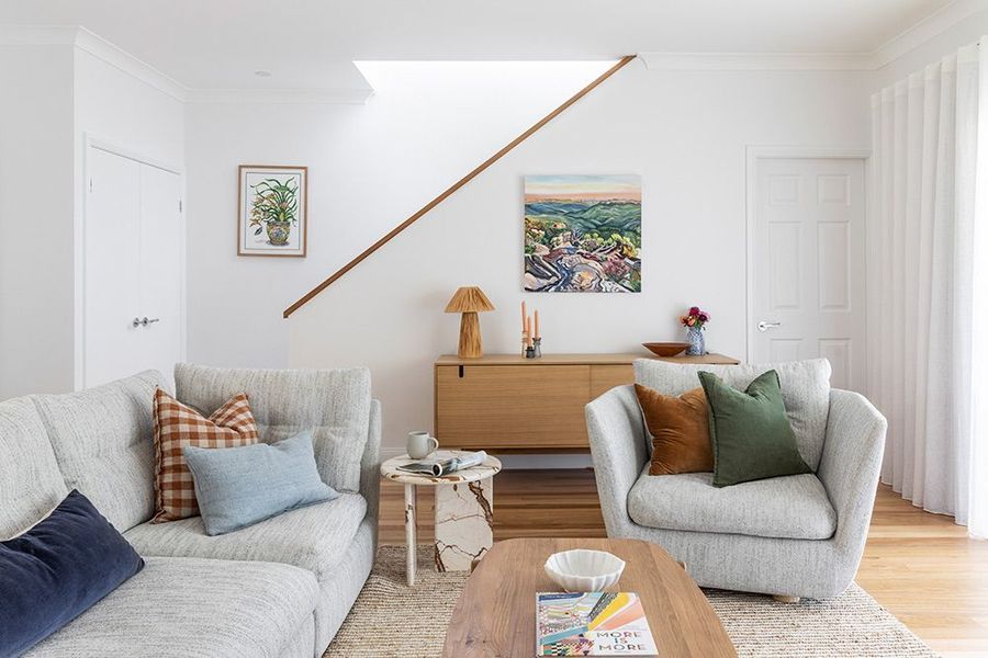 Cozy living room with light-colored sofa, armchair, wooden cabinet, and staircase, painted in white.