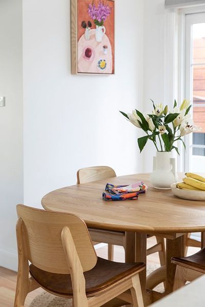Round wooden dining table with chairs, still-life painting on the wall, and vase of lilies.