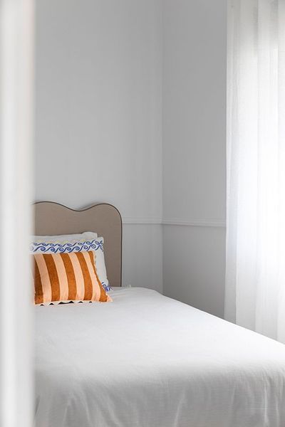 Bed in a white room with striped orange pillow and sheer curtains.