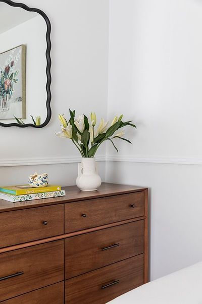 Wooden dresser with flowers in a white vase, beneath a scalloped mirror and artwork, in a white-walled room.