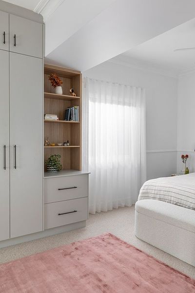 A light-filled bedroom with gray wardrobe, open shelves, pink rug, and white sheer curtains.