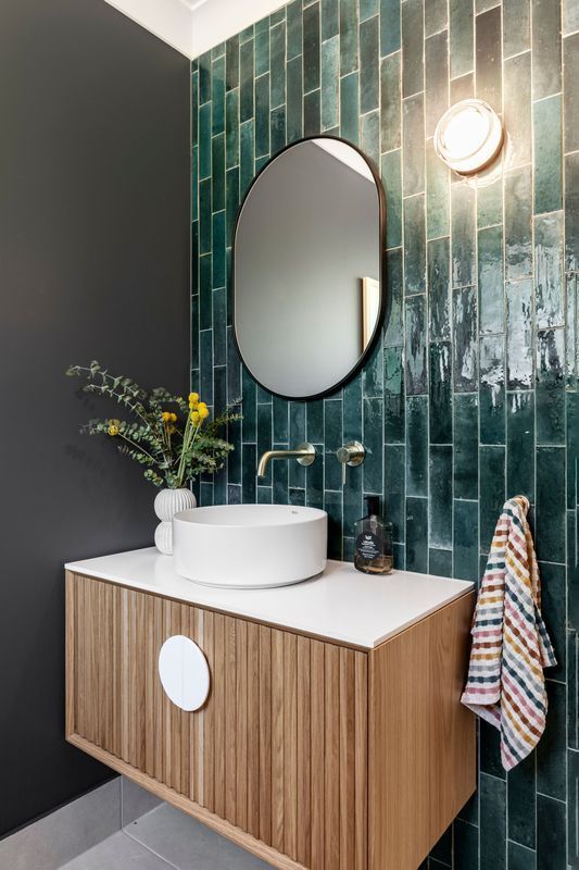 A modern bathroom with a wooden vanity, a green tiled wall, and a dark mirror.