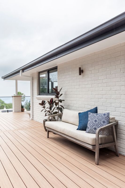 Wooden deck with a light-colored sofa, blue pillows, and white brick wall under a cloudy sky.