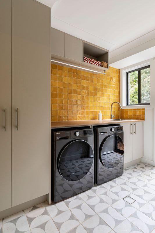 Laundry room with black washer/dryer, yellow tiled backsplash, gray cabinets, and patterned flooring.