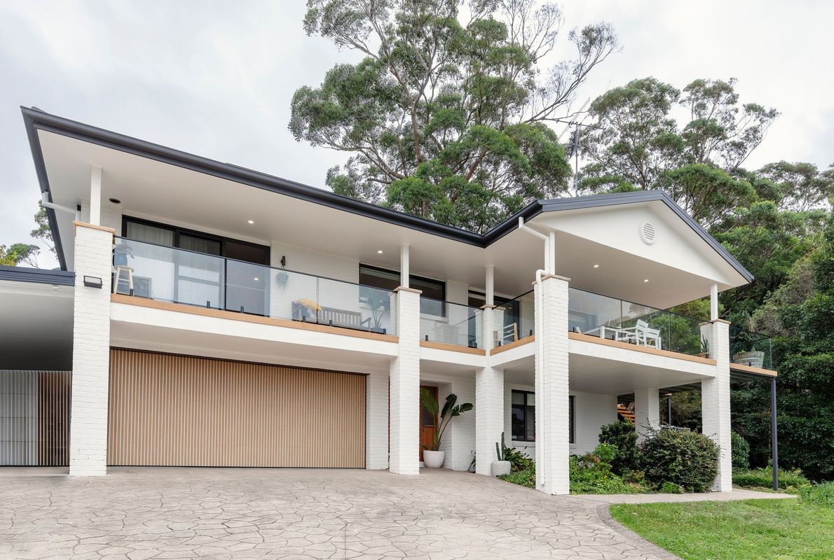 Two-story white house with balconies and a wooden garage door, surrounded by greenery.
