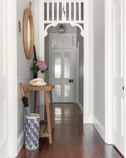 Narrow hallway with wooden floors, white walls, and doorway arch. Wooden table with flowers and mirror on wall.