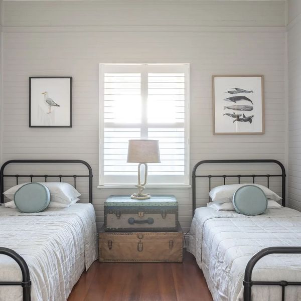 Two beds in a room with light walls, a window, and artwork; a lamp sits on stacked trunks.