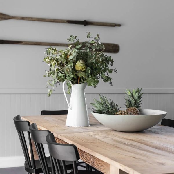 Wooden dining table set with a white vase of greenery and a bowl of pineapples. Black chairs and oars on wall.