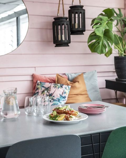 Restaurant table setting with meal, pillows on pink wall, lanterns, and potted plant.