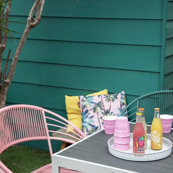 Pink chairs and table with drinks and pillows in a green-walled outdoor setting.