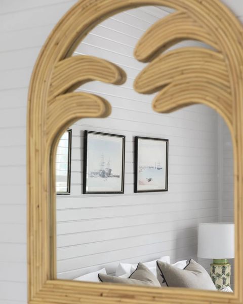 Rattan palm tree mirror reflecting framed art, pillows, a lamp, and a white-paneled wall.