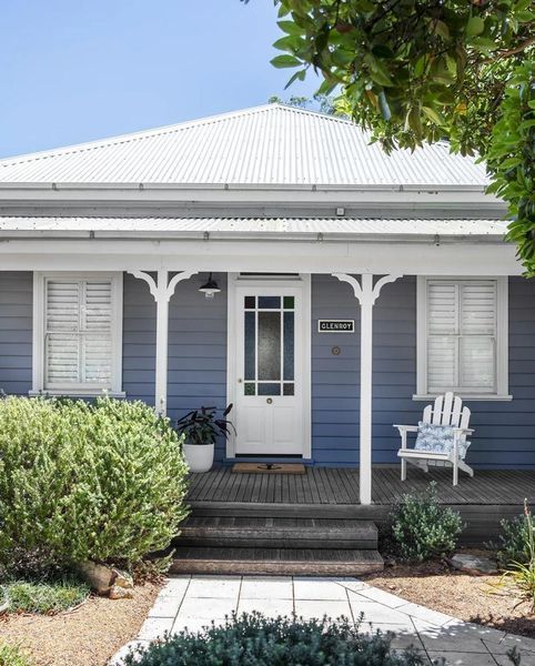 Blue cottage with white trim, porch, and shutters.  A white chair sits to the right of the door.