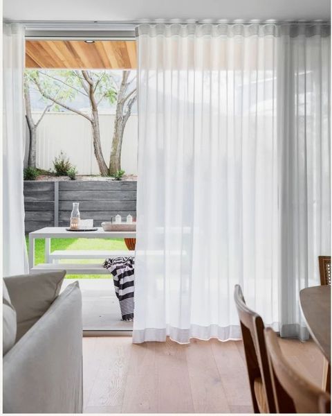 White sheer curtains in front of a glass door open to an outdoor dining area with a table, greenery, and wooden ceiling.