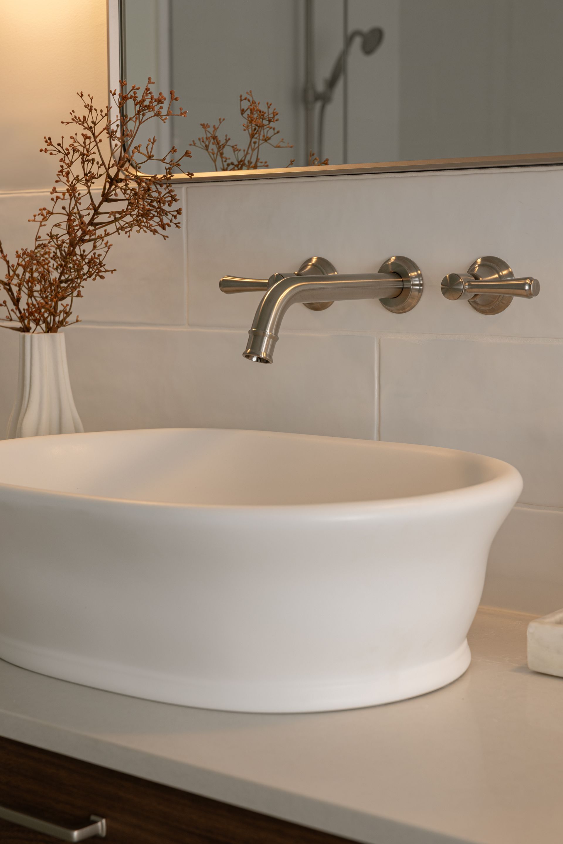 White bathroom sink with wall-mounted faucet. Dried flowers in a vase. White tiled wall.